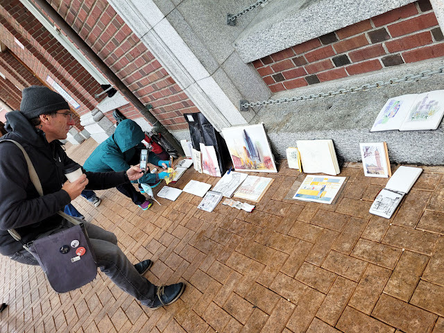 urban sketchers showing each other art works in small notebooks on the ground.